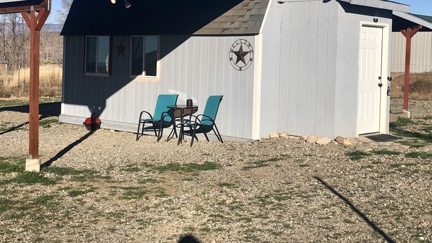 A small, single-story building with a covered porch, a welcome sign, and two chairs. - Tiny House Farm Stay w/Kitchenette (Hotchkiss)