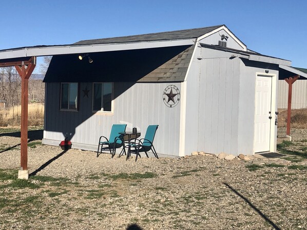 A small, single-story building with a covered porch, a welcome sign, and two chairs. - Tiny House Farm Stay w/Kitchenette (Hotchkiss)