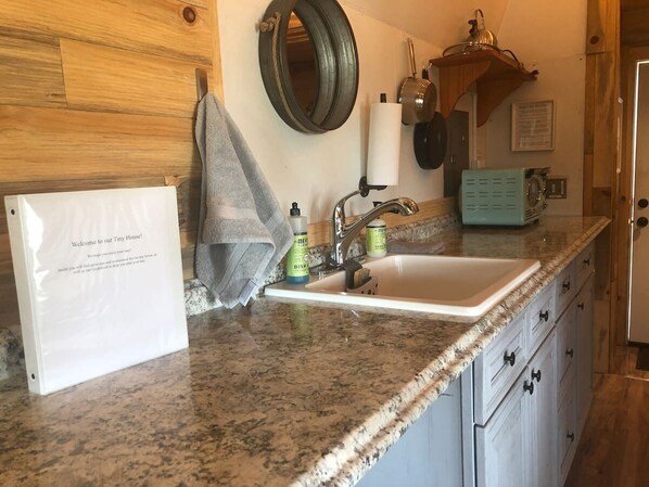 A kitchen with a granite countertop, a sink, a towel, a dish soap dispenser, and a metal pot hanging on the wall. - Tiny House Farm Stay w/Kitchenette (Hotchkiss)