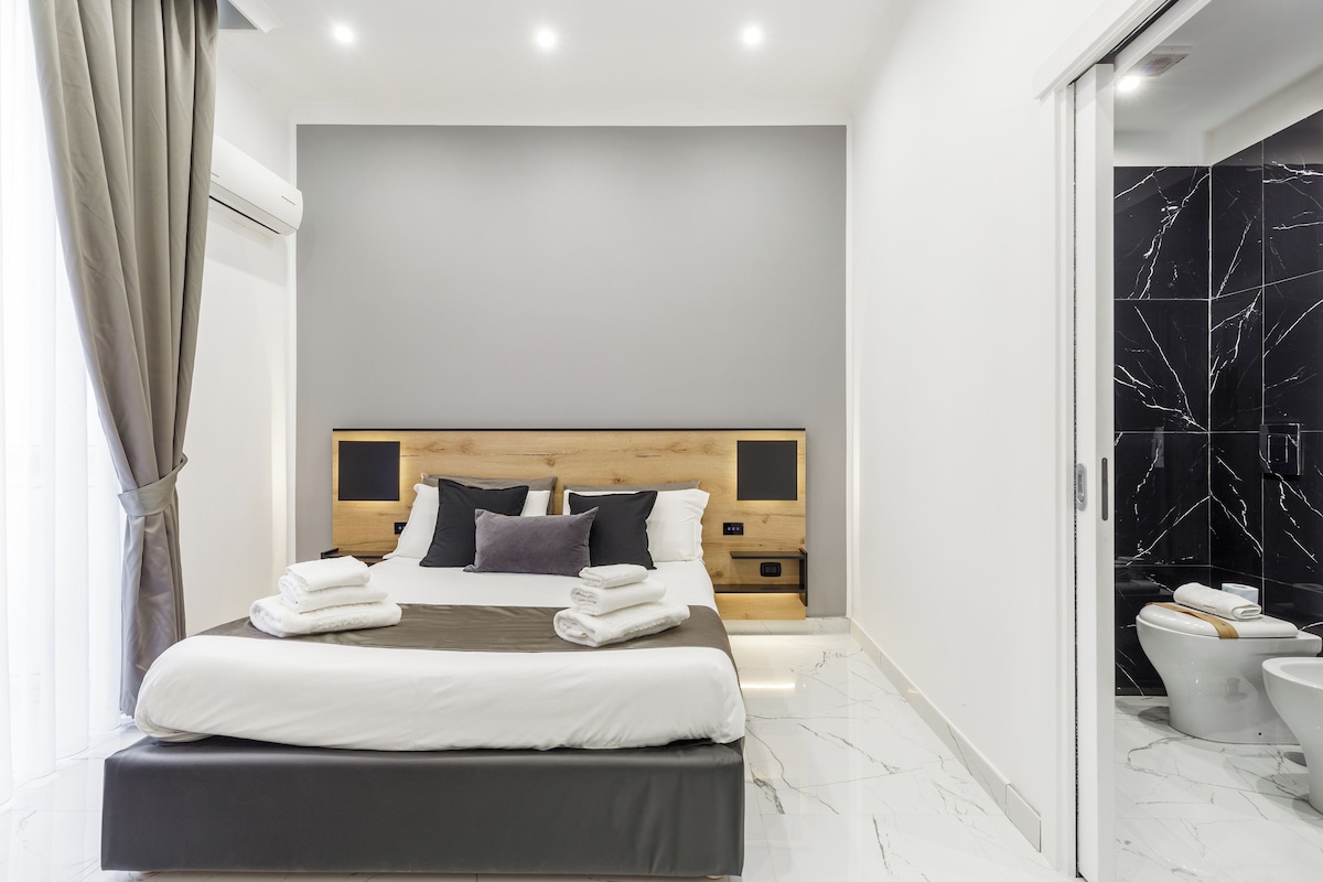 A modern hotel room with a large bed, wooden headboard, and a bathroom with a black marble wall.