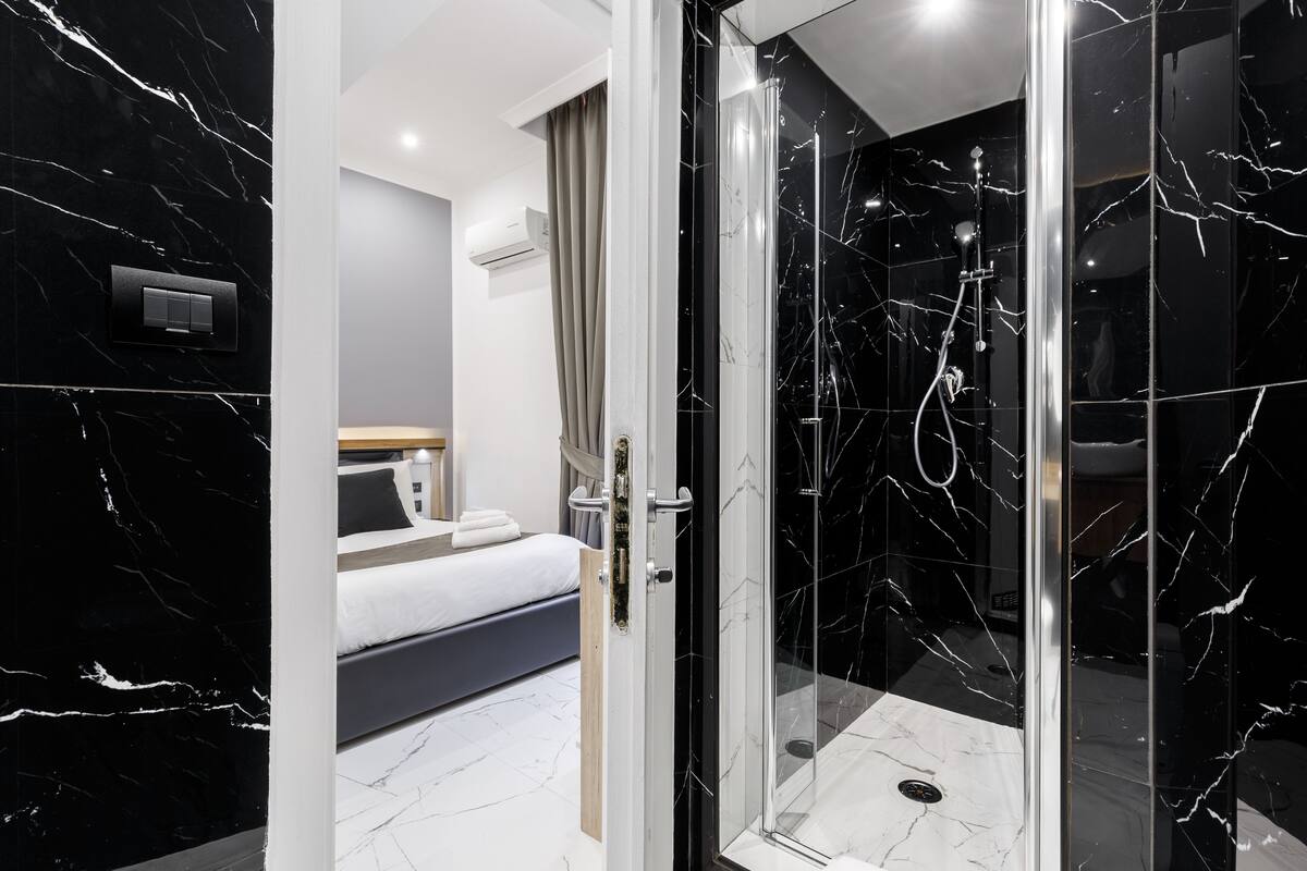 A modern bathroom with a glass-enclosed shower, a bed with a white mattress and gray bedding, and a marble-tiled wall.
