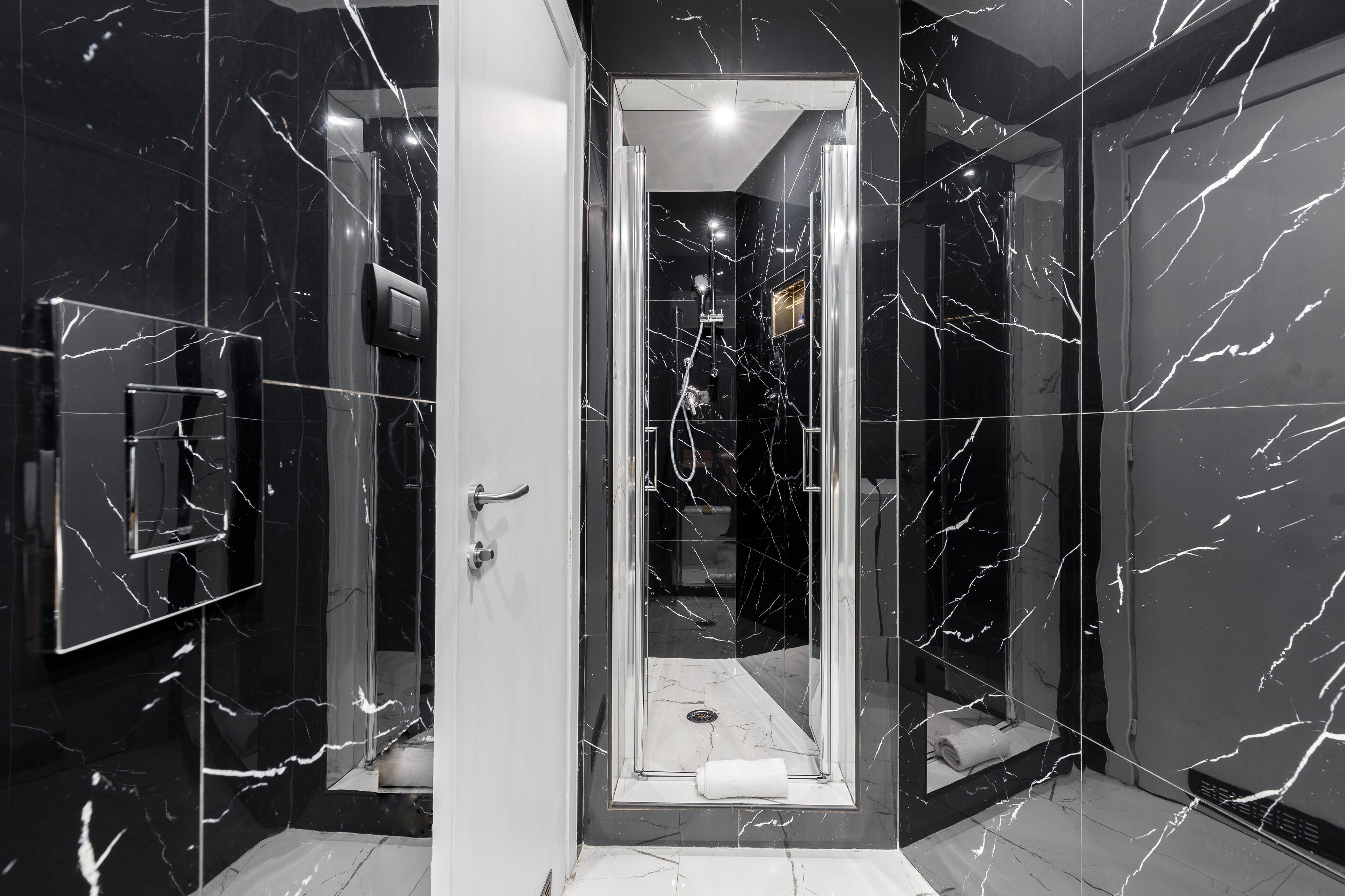 Un bagno moderno con pareti in marmo nero, una cabina doccia in vetro e un asciugamano sul pavimento.