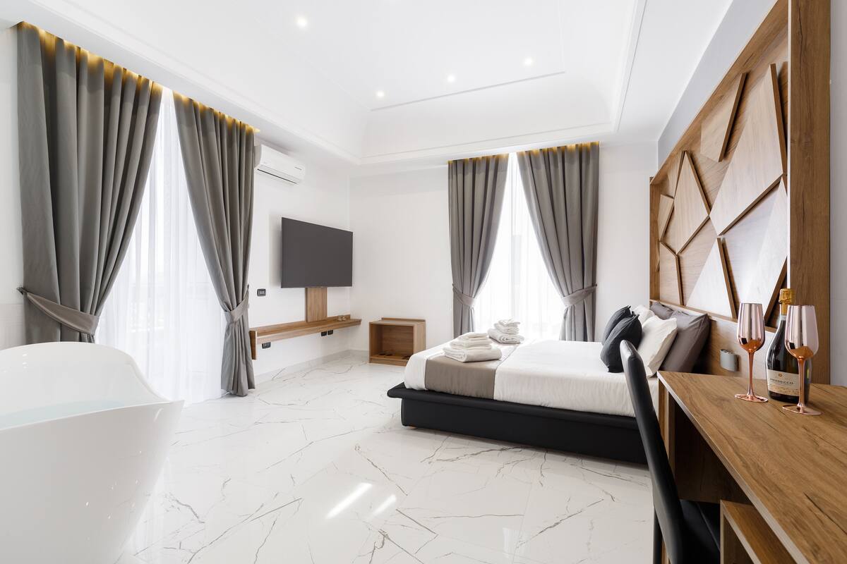 A modern hotel room with a large bed, a wooden desk, a flat-screen TV, a bathtub, and marble flooring.