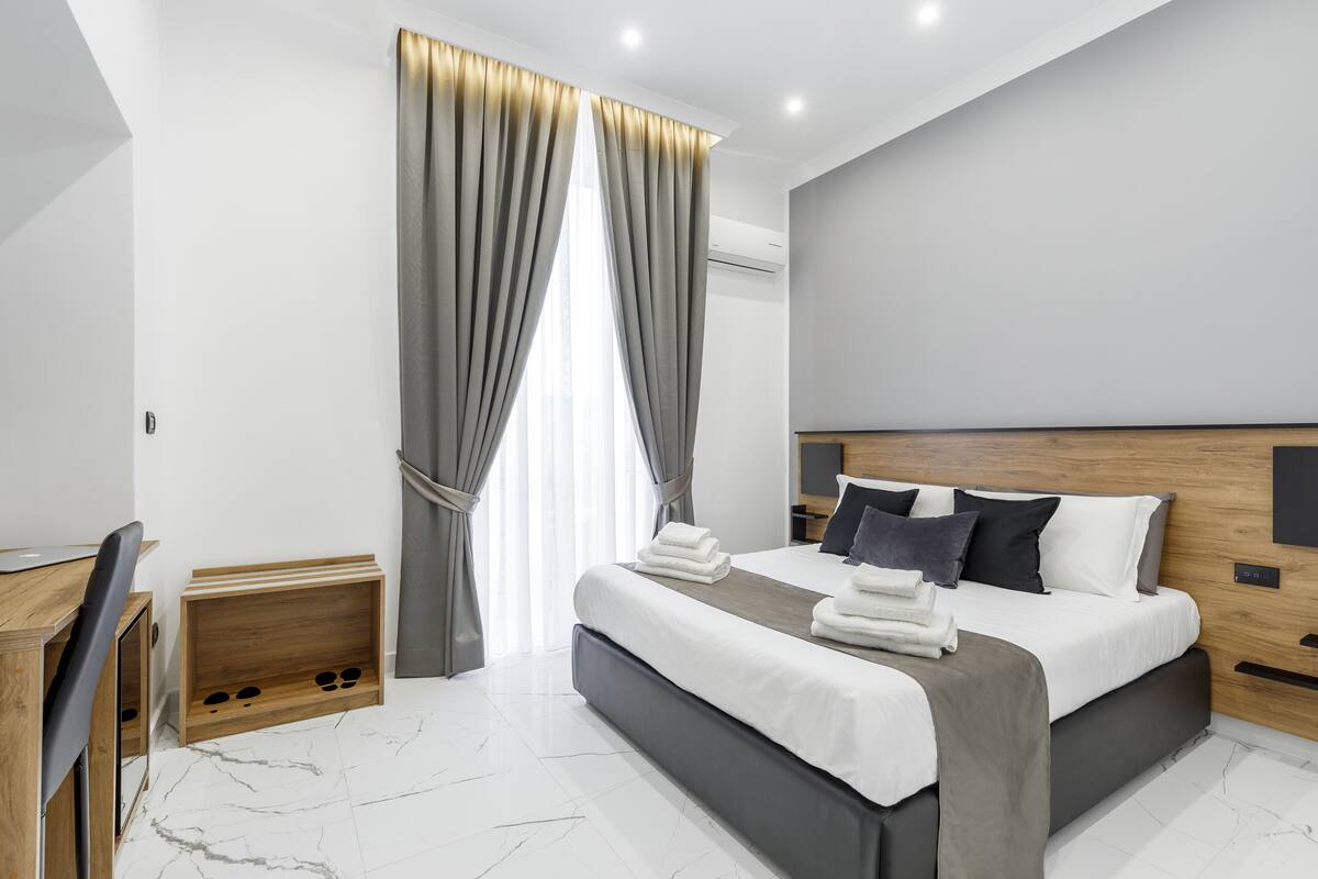 A modern hotel room with a large bed, a wooden headboard, a desk, and a shoe rack.