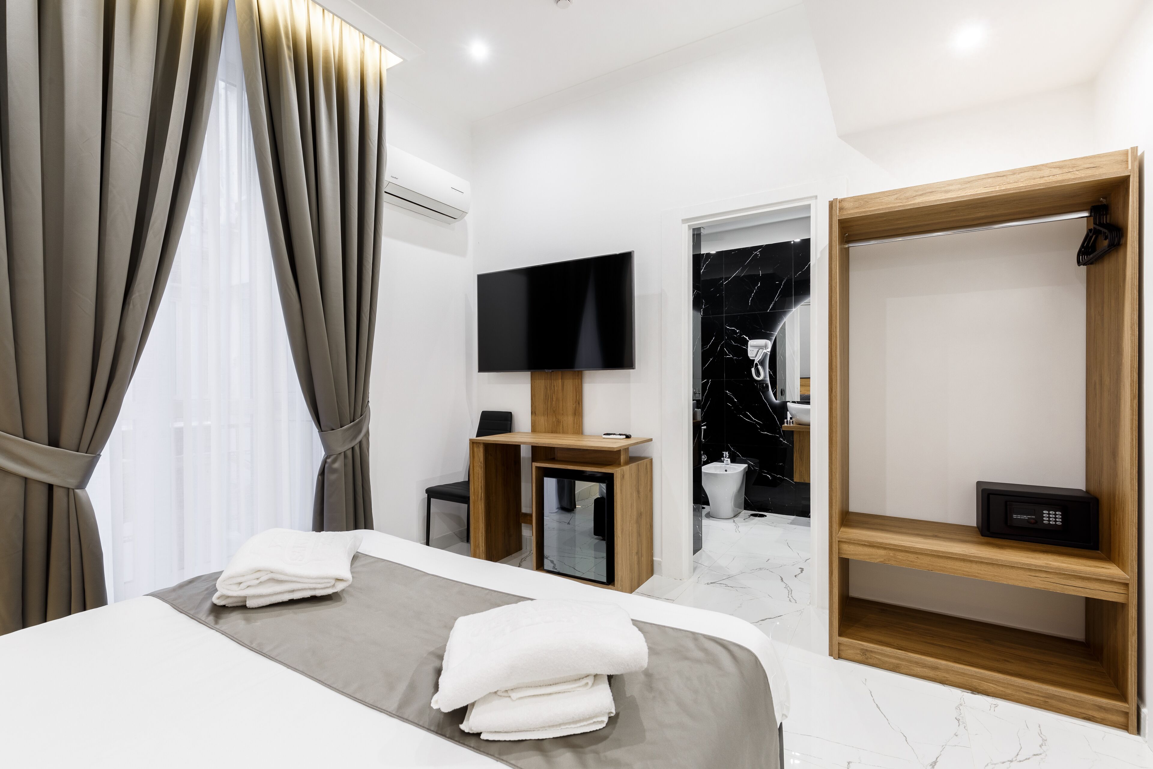 A modern hotel room with a bed, a TV, a wooden shelf, and a bathroom visible through an open door.