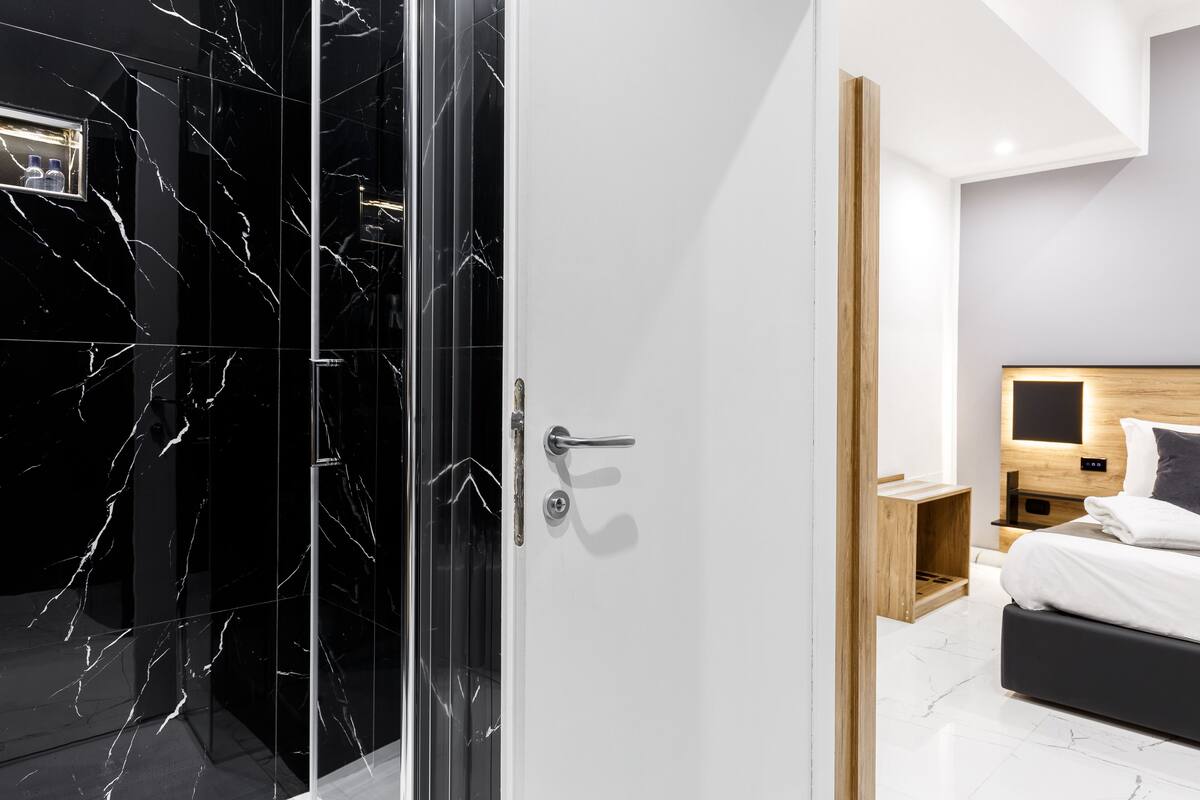 A modern hotel room with a glass shower door, a wooden bedside table, a bed with white bedding, and a headboard with a built-in TV.