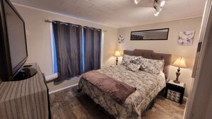 A bedroom with a bed, two bedside lamps, a flat-screen TV, a window with curtains, and a mirror on the wall.