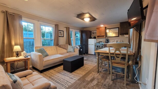 A compact living space with a sofa, coffee table, dining table, and chairs. There is a kitchen area with cabinets and a refrigerator.