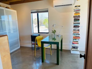 Une pièce avec une table en verre, une chaise jaune, une bibliothèque et une fenêtre donnant sur un beau paysage.