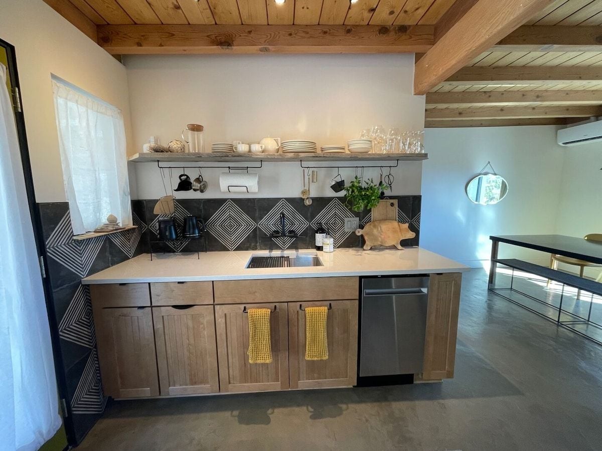 Una cocina con encimera de madera, un fregadero y lavavajillas. Hay armarios debajo de la encimera y una ventana con cortina.