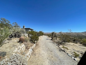 Un chemin de gravier mène à une maison dotée d’une cheminée, qui est entourée de végétation désertique et surplombée par un ciel bleu clair.