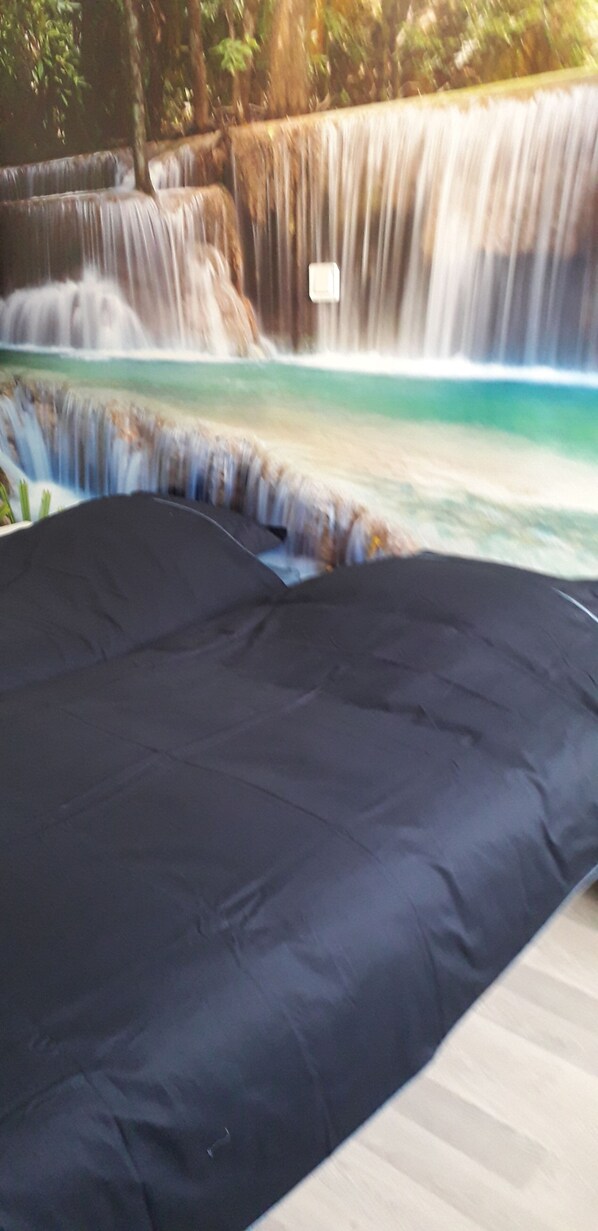 Una cama con un cubre oscuro en una habitación con vistas a una cascada.