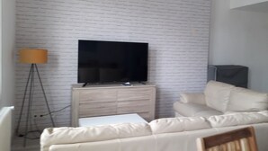 Un salon avec un téléviseur à écran plat posé sur un meuble en bois, un canapé beige, une petite table blanche et un lampadaire.