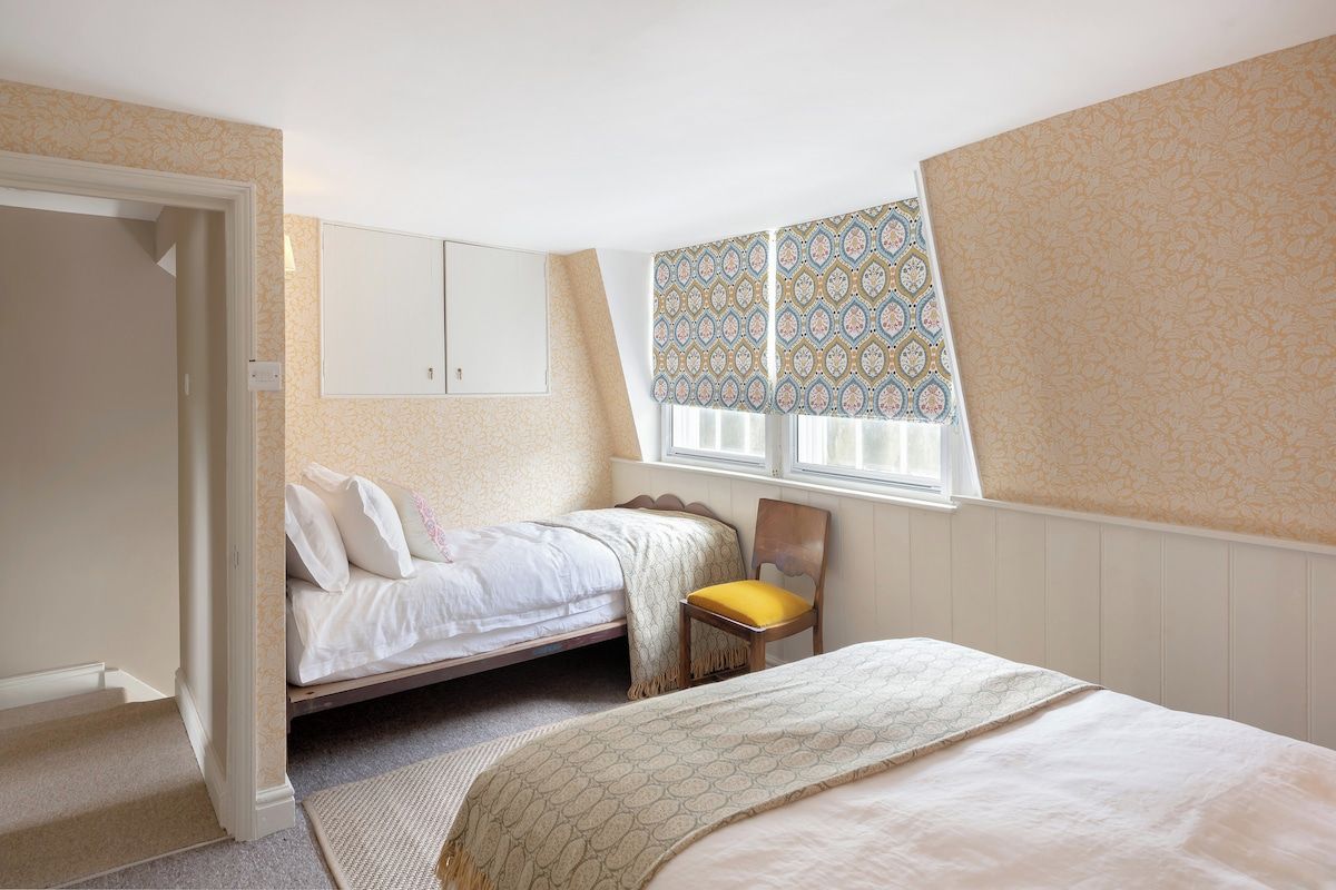 A bedroom with a single bed, a chair, a wardrobe, and a window with patterned curtains.