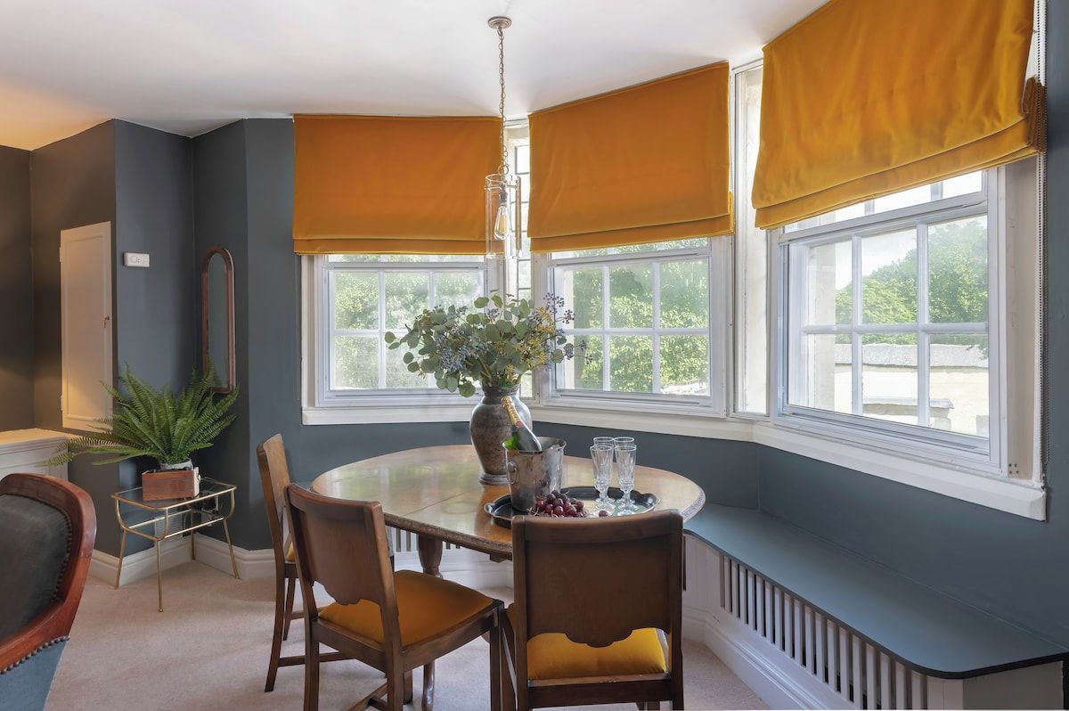 Un coin repas avec une table ronde, des chaises, un vase de fleurs et une fenêtre avec des stores.