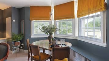 A dining area with a round table, chairs, a vase with flowers, and a window with blinds.