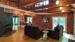 Uma sala de estar ampla com teto de madeira, lareira, sofá, televisão e área de jantar com cadeiras.