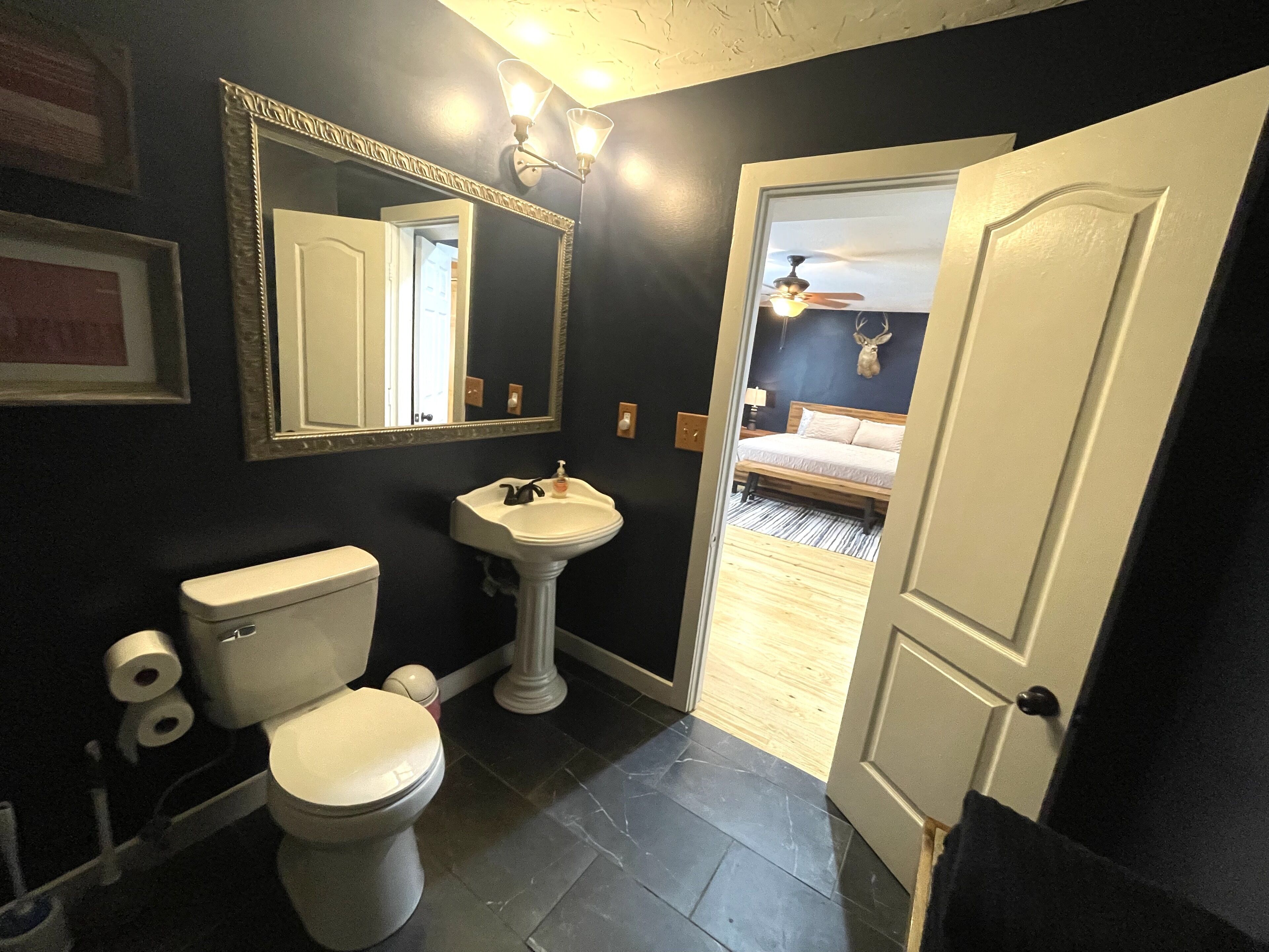A bathroom with a toilet, sink, and mirror. There is a door leading to a bedroom with a bed and a ceiling fan.