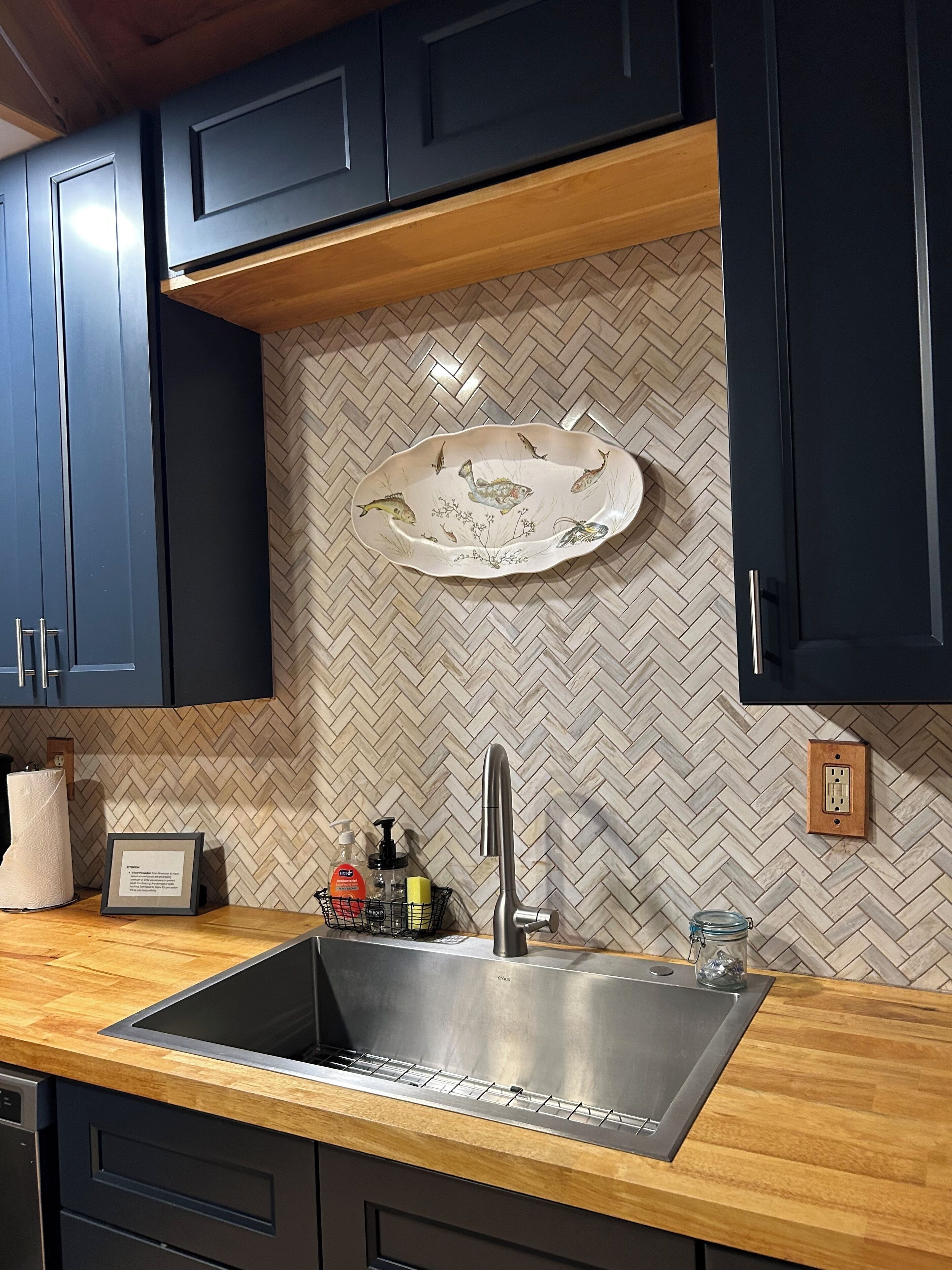 Een keuken met donkere kastjes, een backsplash met een geblokt patroon, een roestvrijstalen spoelbak en een houten aanrecht.