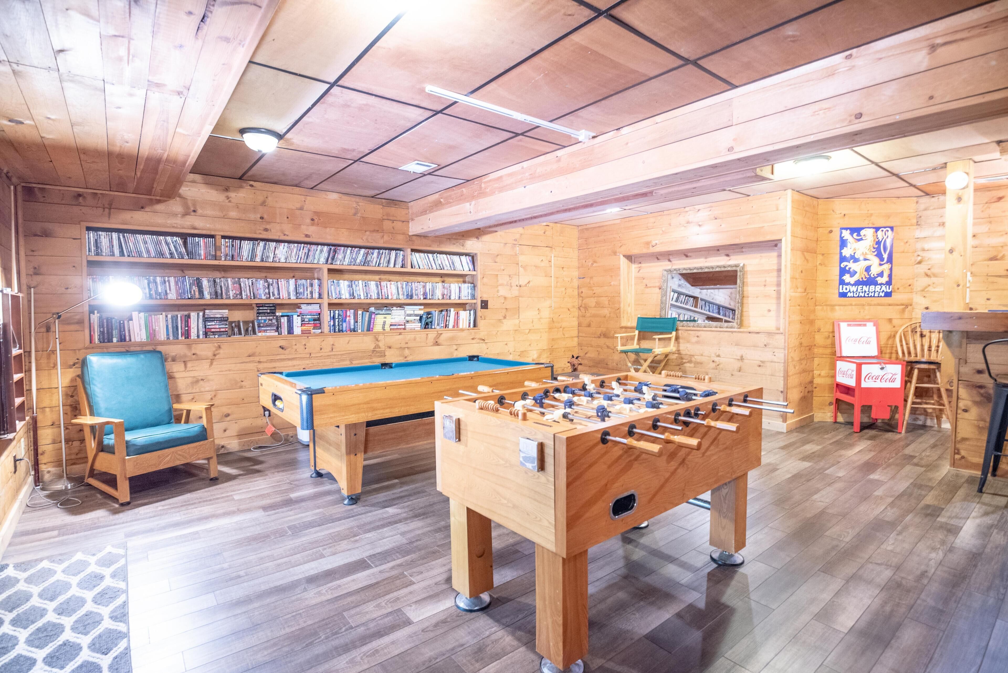 A wooden-floored room with a foosball table, a pool table, and a bookshelf filled with DVDs.