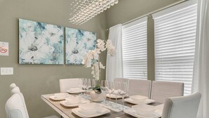 Comedor con una mesa rectangular dispuesta para seis comensales, sillas blancas y dos pinturas abstractas de flores en la pared.