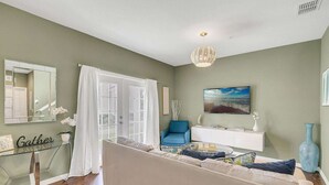 Una sala de estar con un sofá color beige, una mesa de centro de vidrio, un sillón azul, una consola blanca y un televisor de pantalla plana grande montado en la pared.