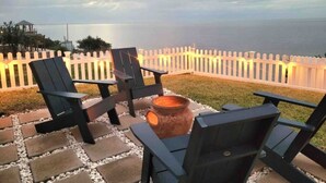 Un espacio al aire libre con sillas Adirondack y un pozo de fuego, con vistas a un cuerpo de agua y una cerca blanca de piquetes.