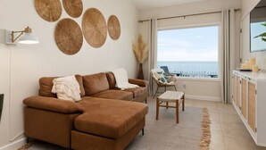 Una sala de estar moderna con un sofá marrón, un sillón pequeño y vista al mar a través de grandes ventanales.