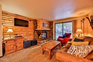 Interior de una cabaña de troncos con chimenea de piedra, un televisor de pantalla plana en la pared y una zona de estar acogedora con un sillón rojo y un sofá marrón.