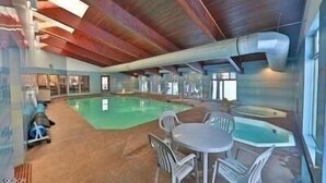 Piscina interior con techo de madera, paredes de vidrio transparente y una zona de estar con sillas.