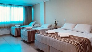 Habitación de hotel con tres camas, una planta en maceta y un amplio ventanal con cortinas.