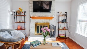 Una sala de estar con chimenea de ladrillo blanco, un televisor de pantalla plana montado encima y una zona de estar acogedora con una silla de mimbre y una mesa de centro.