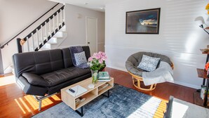 Una sala de estar moderna con un sofá de cuero negro, una mesa de centro de madera, un sillón gris y una escalera al fondo.
