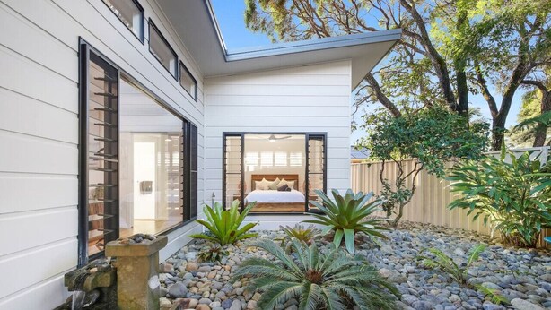 Una casa moderna con un dormitorio visible, un elemento decorativo de piedra con agua y un jardín con diversas plantas.