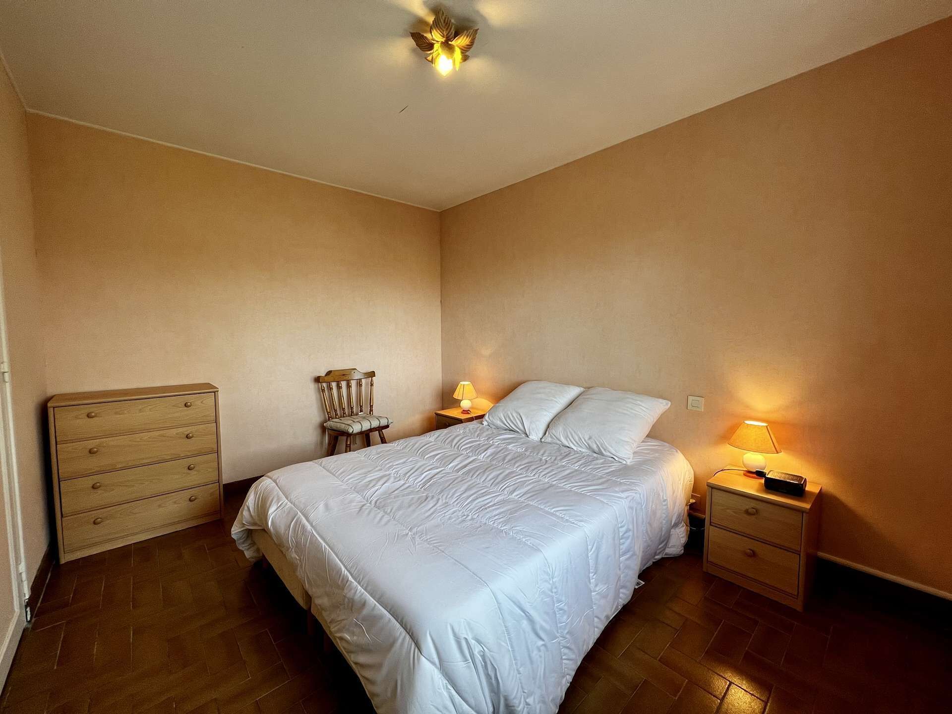 Ein Schlafzimmer mit einem Bett, einem hölzernen Schminktisch, einem Sessel, zwei Nachttischen mit Lampen und einer Deckenleuchte.