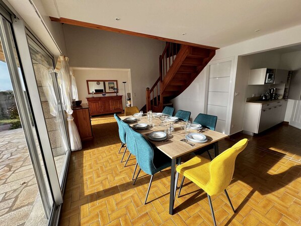Un coin repas avec une table en bois dressée pour quatre personnes, des chaises bleues et jaunes, et une cuisine en arrière-plan.