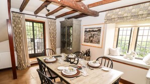 A dining room with a wooden table set for four, black chairs, a cabinet, and a window with a view of greenery. - The Small House (Cheltenham)