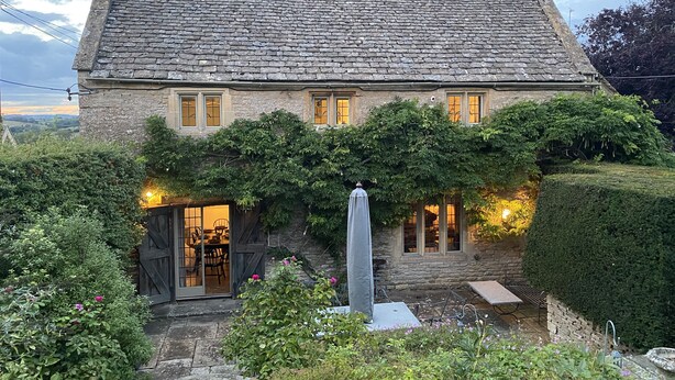 A stone house with a sloped roof, a covered porch, and a garden area with a table and chairs. - The Small House (Cheltenham)