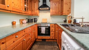 A compact kitchen with wooden cabinets, a built-in oven, a washing machine, and a sink. - Beckfold Cottage (Broughton in Furness)