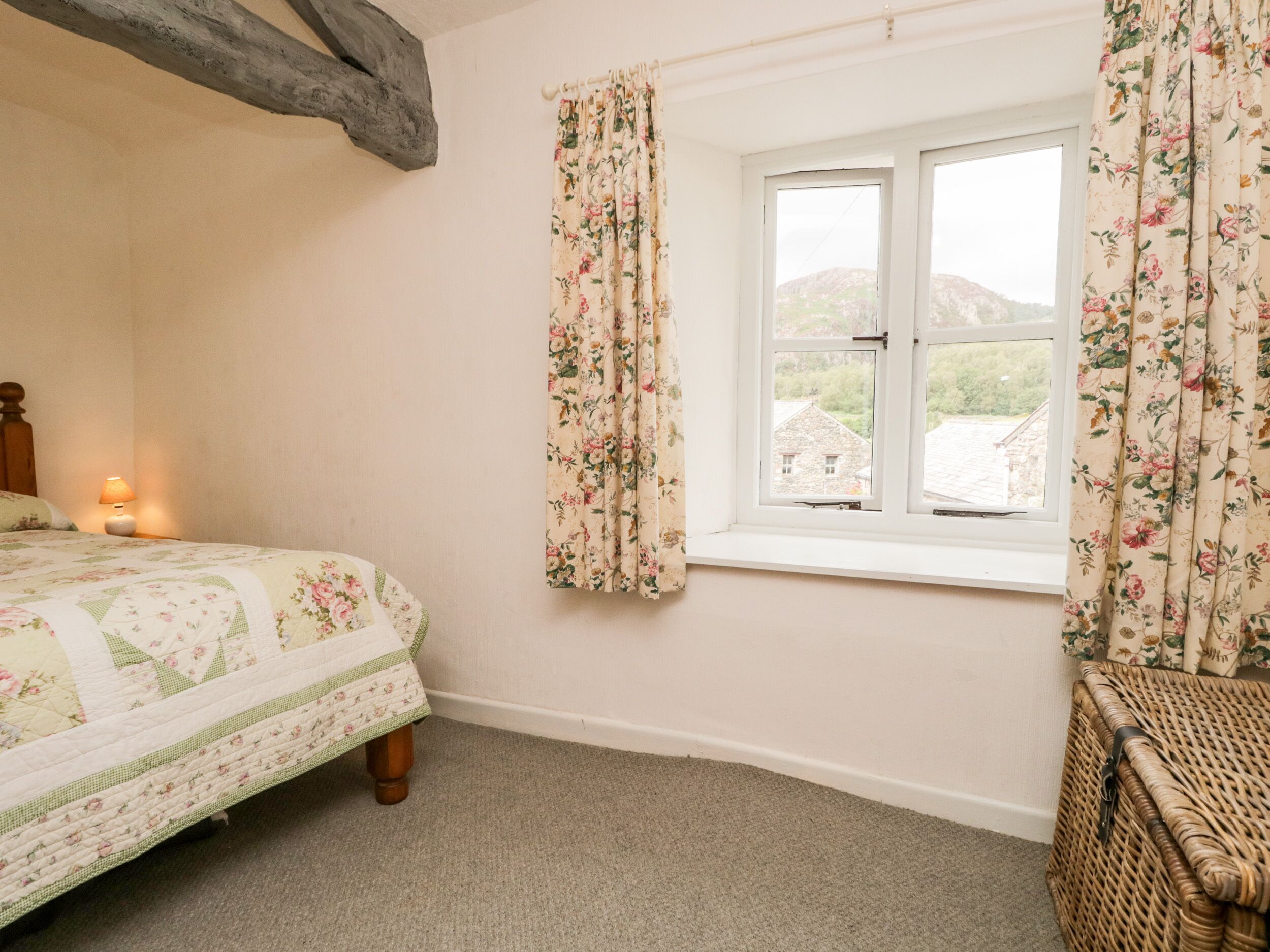 Un dormitorio con cama, ventana con cortinas florales, lámpara y una silla de mimbre.