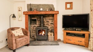 A cozy living room with a stone fireplace, a comfortable armchair, a flat-screen TV, and a wooden entertainment unit. - Beckfold Cottage (Broughton in Furness)