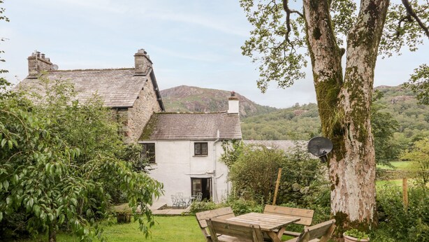 A rustic house with a stone chimney, a wooden table and chairs in the yard, and a large tree with a satellite dish. - Beckfold Cottage (Broughton in Furness)