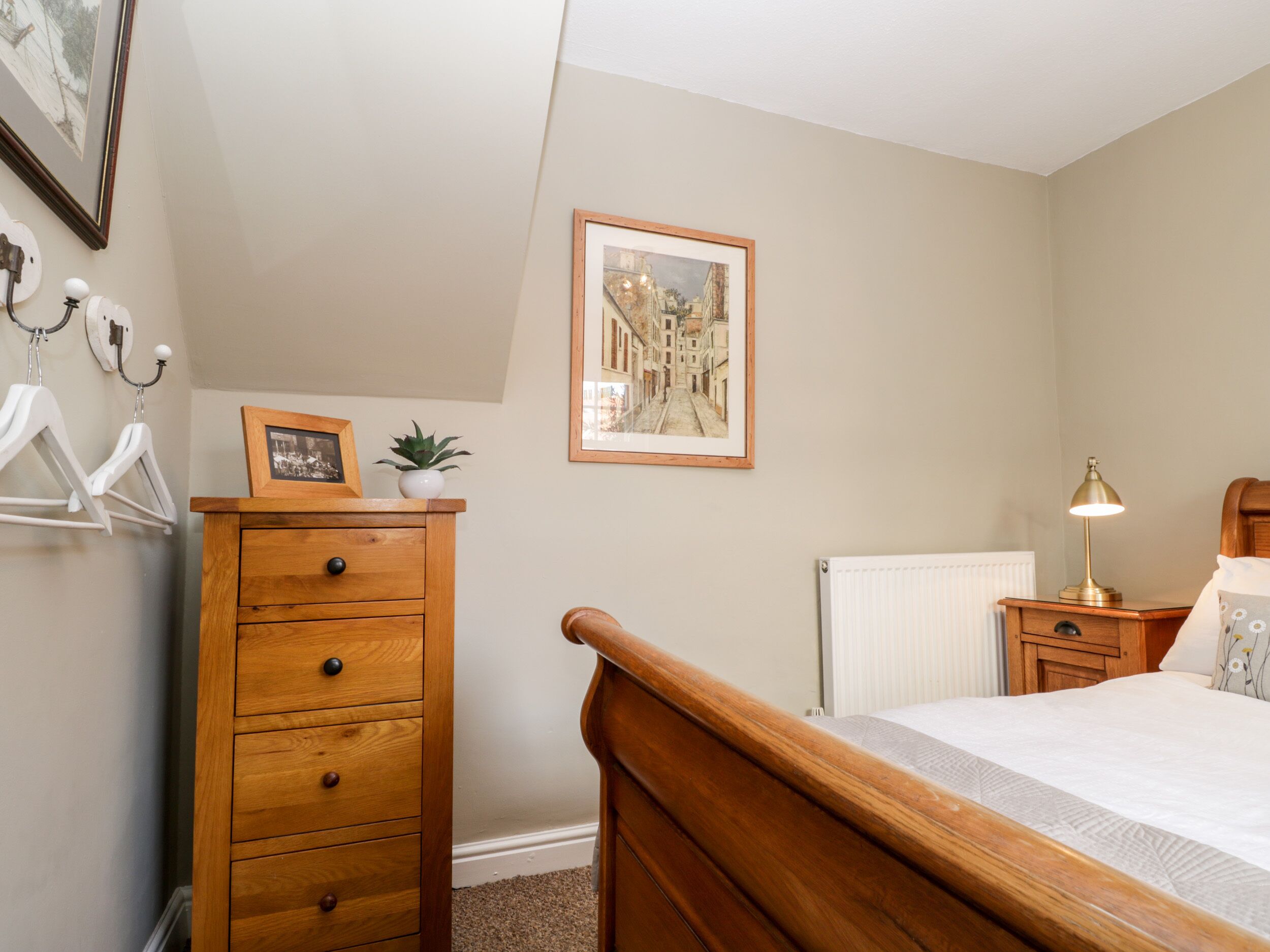 A bedroom with a wooden dresser, a bed, a nightstand with a lamp, a radiator, and a framed picture on the wall.