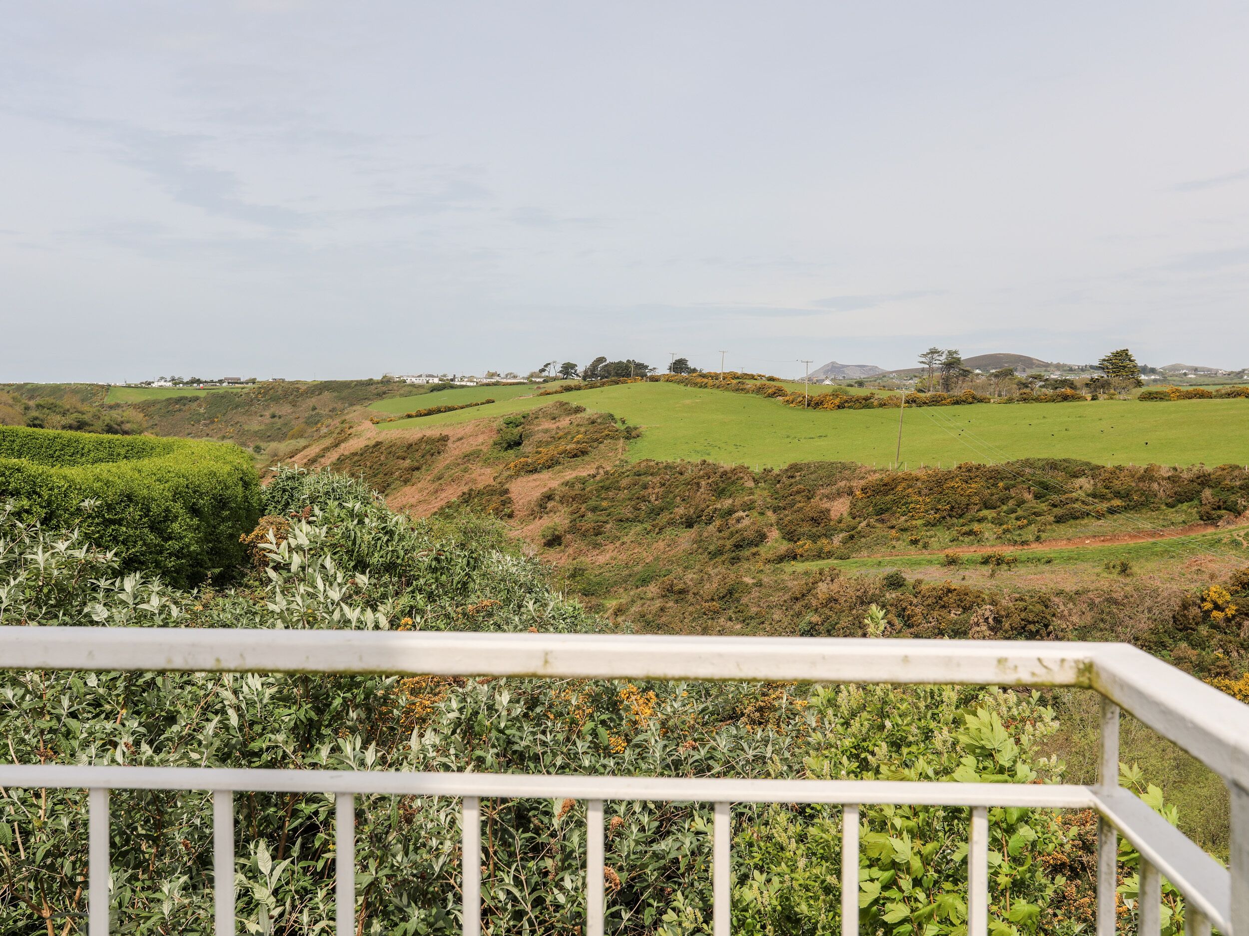 Blick von einem Balkon mit weißer Brüstung auf eine grüne Landschaft mit sanften Hügeln, vereinzelten Bäumen und einigen Gebäuden in der Ferne.