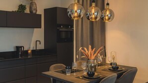 Ein moderner Essbereich mit einem dunkelholzigen Tisch, gedeckt für vier Personen. Stühle umgeben den Tisch. Eine hohe, filigrane Leuchte hängt darüber, und auf dem Tisch steht eine Vase mit Trockenblumen.