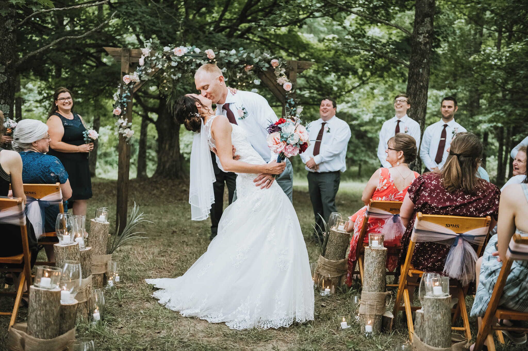 A wedding ceremony outdoors with guests seated on wooden chairs, a floral arch, and candles on stands.