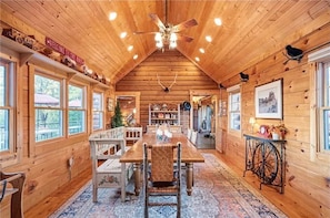 Ein Interieur einer Holzhütte mit einem Esstisch, Stühlen, einem Deckenventilator und einem großen Fenster.