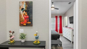 Quarto com pintura de personagem de desenho animado, uma pequena mesa com bonecos e vista para outro quarto com uma cama e uma TV.
