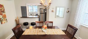 A dining room with a wooden table, four chairs, a chandelier, and a kitchen in the background. - Immaculate Midtown home for Work teams & Families (Augusta)