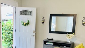 A living room with a sofa, a coffee table with a decorative tray, a door with a wreath, a wall-mounted mirror, and a small cabinet with books.
